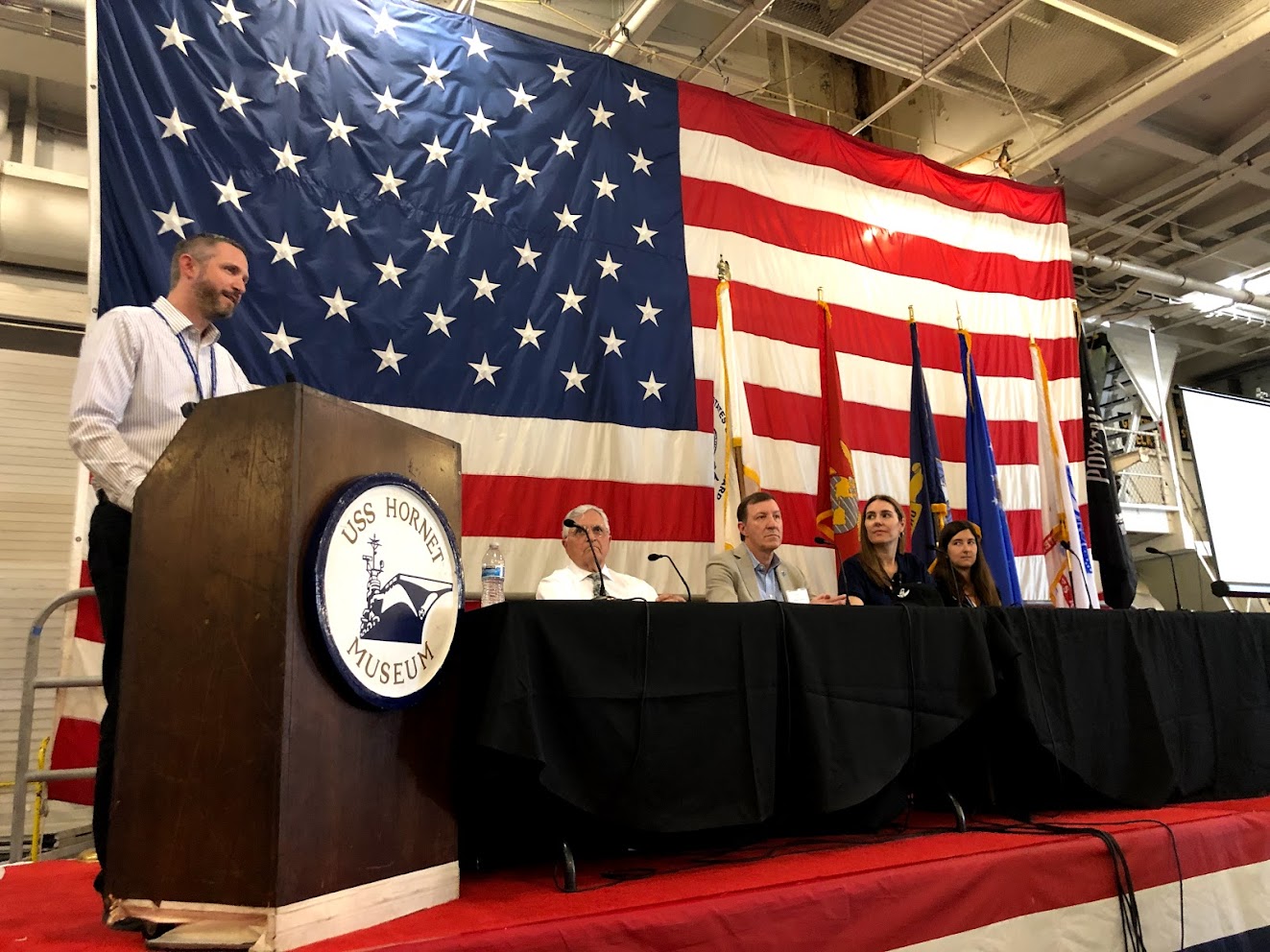 The discussion panel on stage below deck on the Hornet. Jake Bleacher, NASA Chief Exploration Scientist is at the podium and Jack Schmitt, Apollo 17 Lunar Module Pilot, is seated on the panel with other scientists